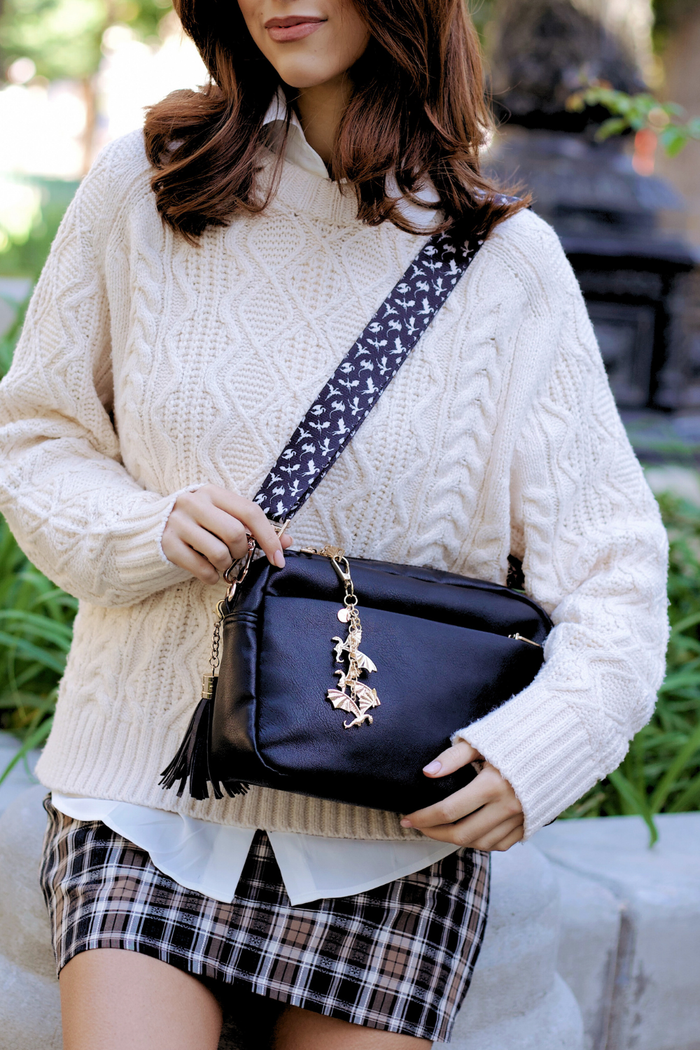 Person wearing a white cable knit sweater and plaid skirt, holding a black handbag with a decorative dragon charm and a black and white dragon bag strap.