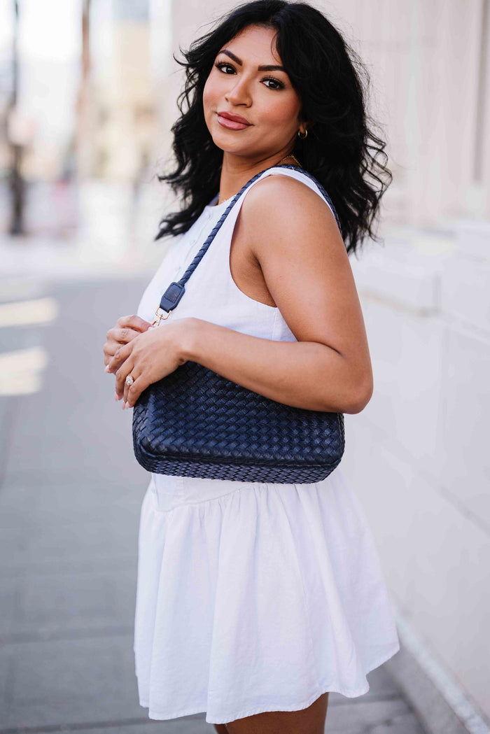 Woman in white dress holding a blue A woman wearing Modern + Chic Elle Woven Crossbody Bag for Women over her shoulder outdoors on a street.