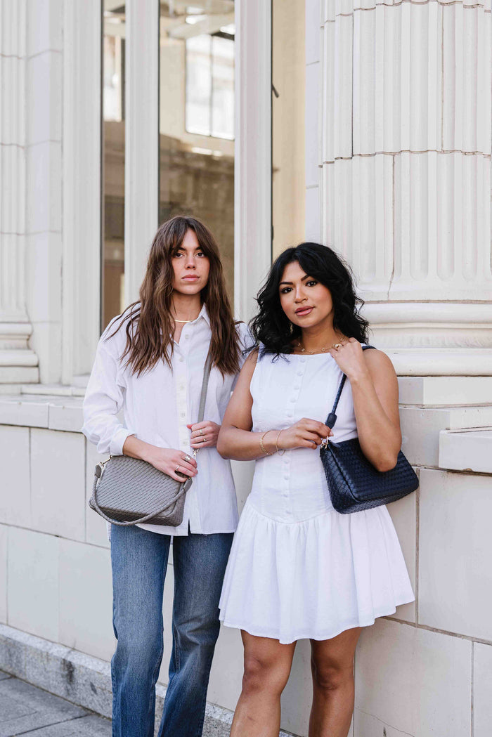 Two women standing next to each other, one holding a gray Modern + Chic Elle Woven Crossbody Bag for Women over crossbody and the other holding a black one. Both are wearing white and the setting appears to be outdoors with columns in the background.