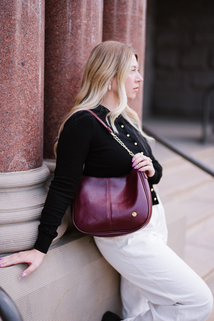 Woman leaning against a wall with a wine Modern + Chic Marissa vegan leather handbag on her should displaying gold chain accents on the strap.