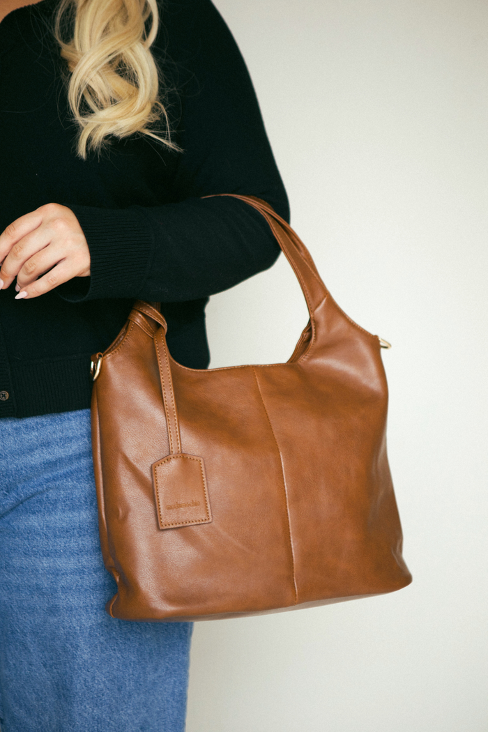 Brown leather handbag held by a person wearing a black sweater and blue jeans on a light background