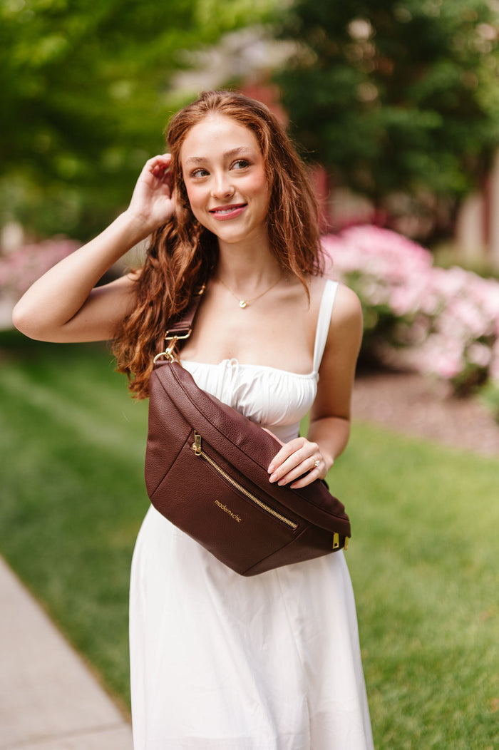 A woman wearing a white dress holding an oversized brown Modern+Chic Presley Oversized Sling Bag for Women with gold hardware as she wears it crossbody.