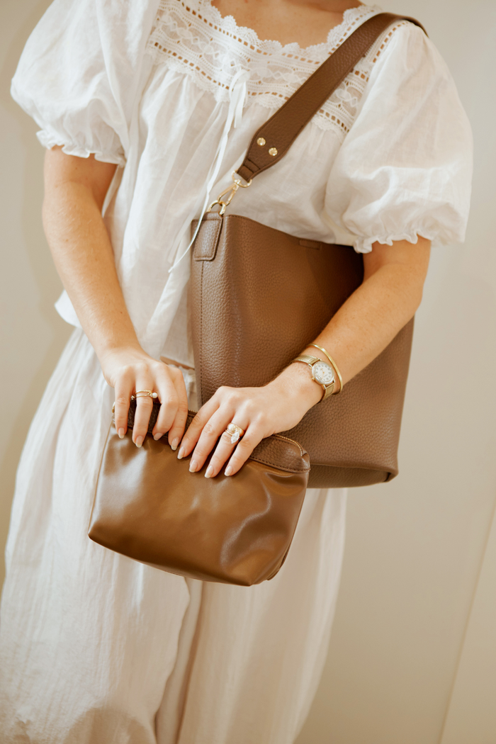 A woman holding a brown Modern+Chic vegan leather Sophie Bucket Bag and coordinating zipper pouch for Women –  with a blurred background.