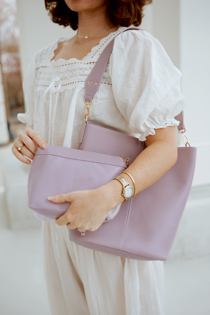 A woman holding a light violet Modern+Chic vegan leather Sophie Bucket Bag and coordinating zipper pouch for Women –  with a blurred background.