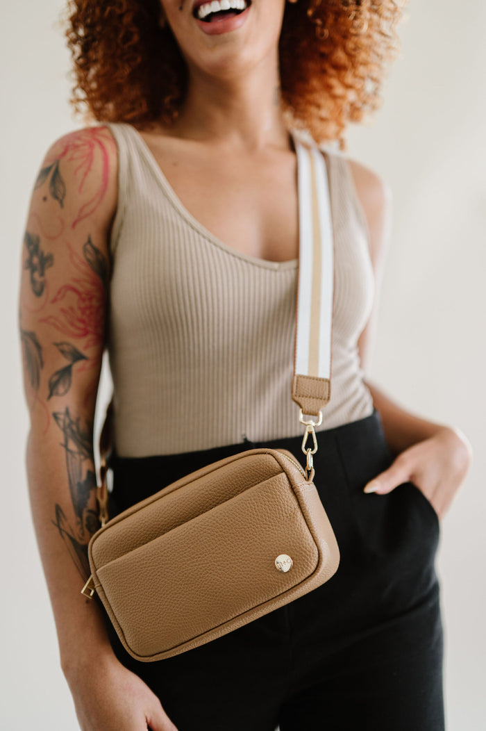 A woman wearing a camel colored Modern+Chic Willow Vegan Leather Convertible Crossbody Bag on a white background.